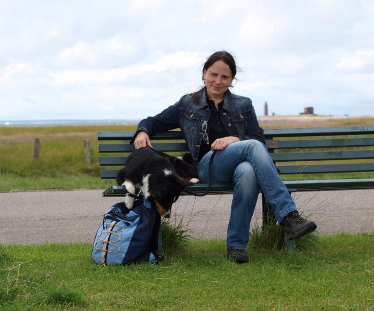 Die Autorin sitzt mit ihrem Hund auf einer Bank, entspannte Landschaft im Hintergrund.